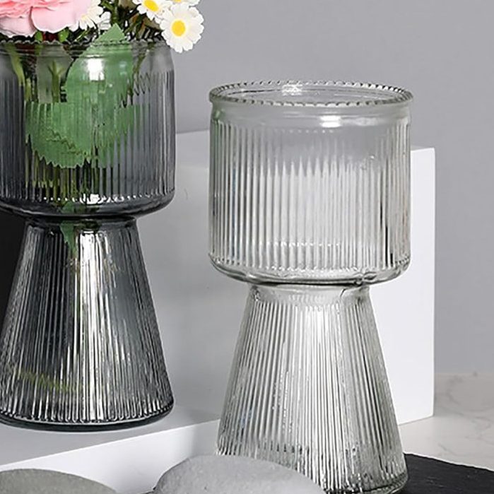 Two ribbed glass vases, one with pink roses and white daisies, are displayed next to smooth gray stones on a white surface against a gray background.