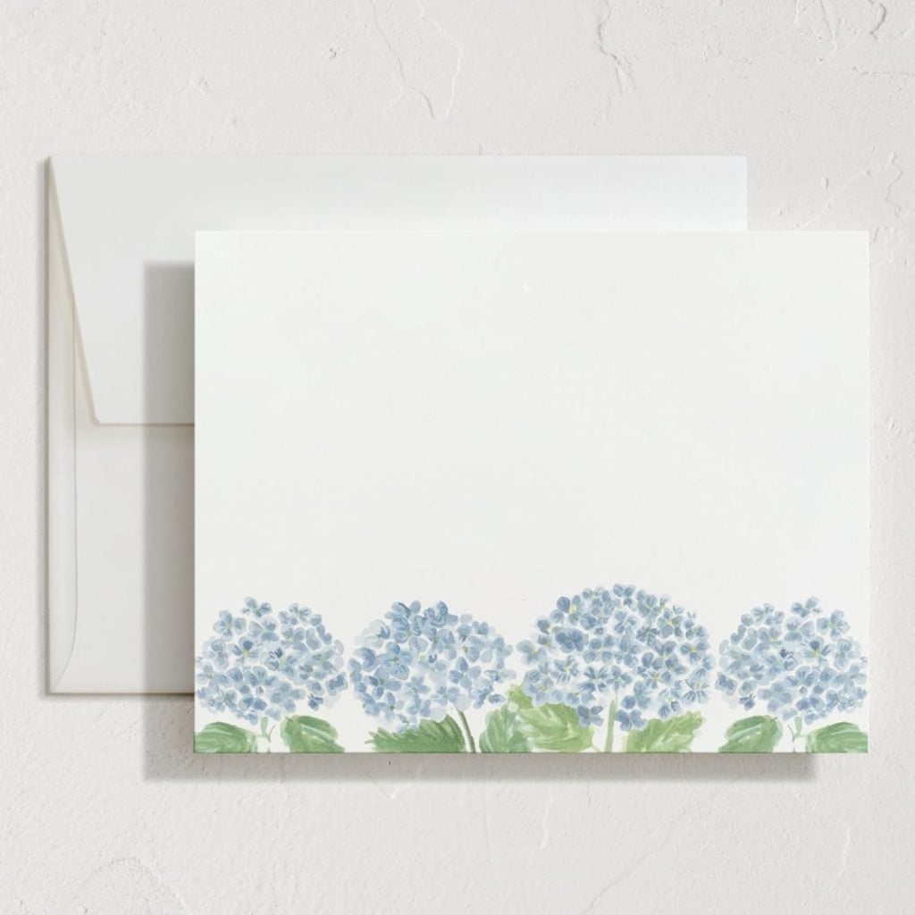 A blank notecard with a row of blue hydrangea flowers and green leaves at the bottom, placed in front of a matching white envelope.
