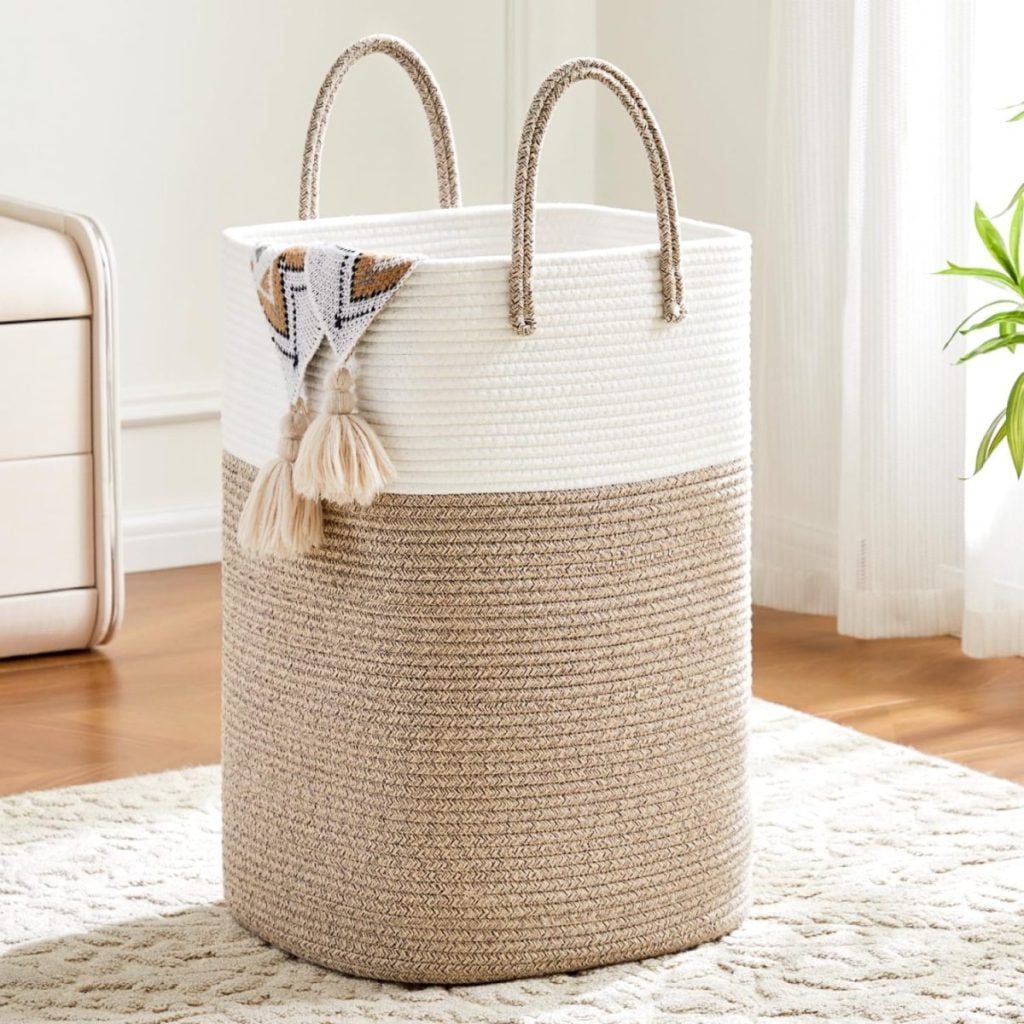 A woven laundry basket with two handles, featuring a white and beige color block design, sits on a textured rug in a bright room. A patterned tassel cloth hangs over the side.