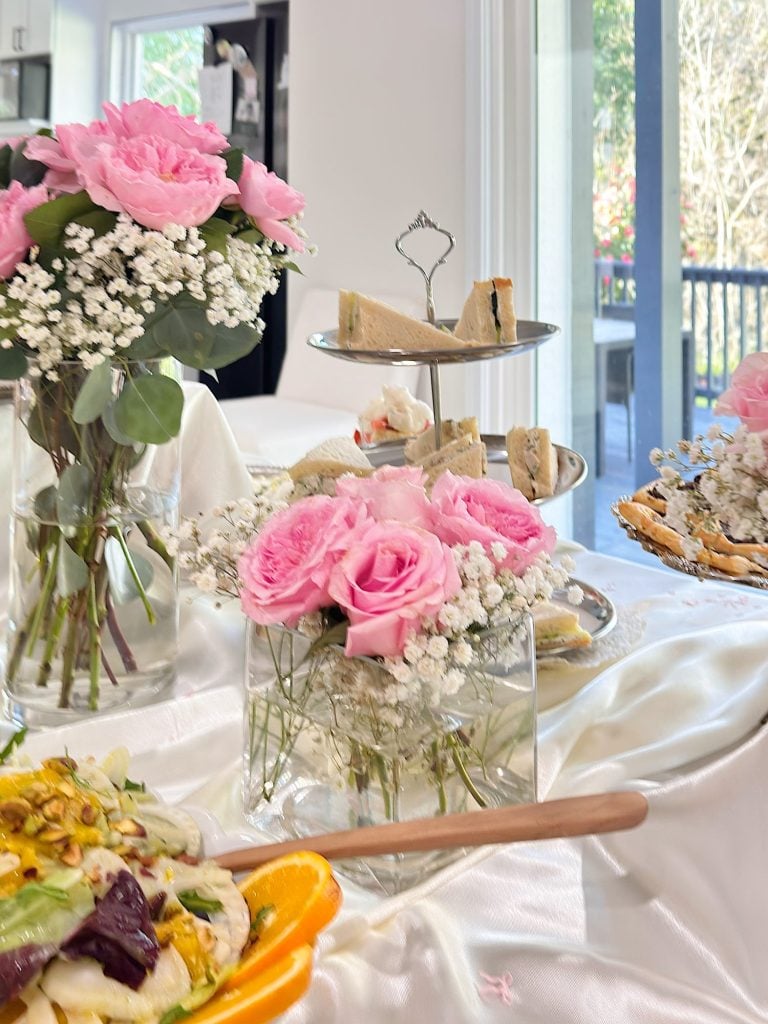 Table set for tea with pink roses, baby's breath, a tiered tray of sandwiches, salad, and a window view of greenery in the background.
