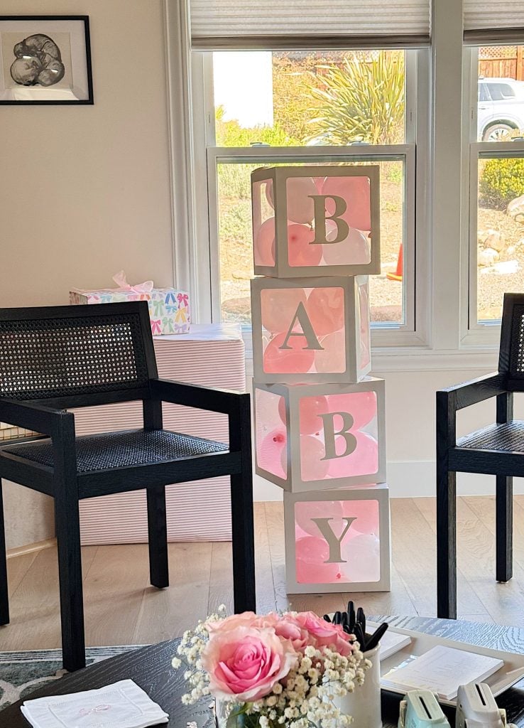 Four stacked boxes with the letters B, A, B, Y stand between two black chairs in a well-lit room decorated for a baby shower. Pink flowers and gifts are visible.