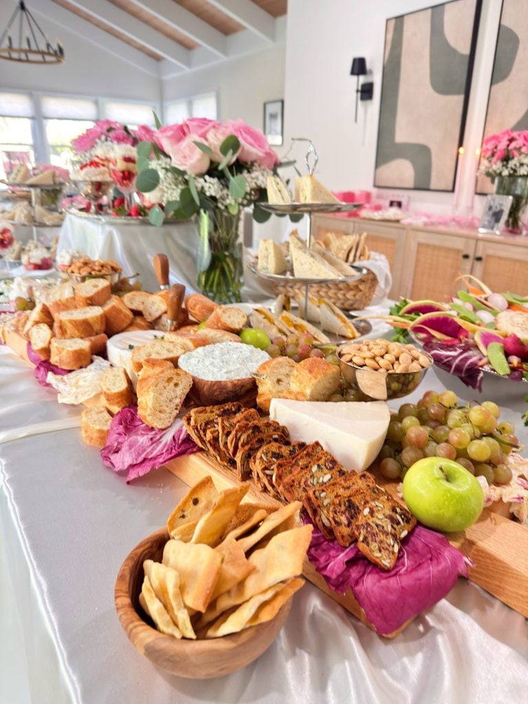 A table set with an assorted charcuterie board featuring cheeses, breads, fruits, crackers, nuts, and dips, surrounded by floral arrangements and serving dishes.