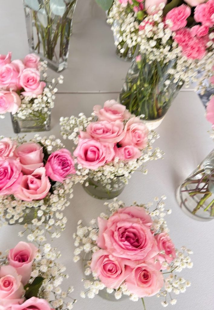 Several glass vases filled with pink roses and white baby's breath flowers are arranged on a light-colored surface.