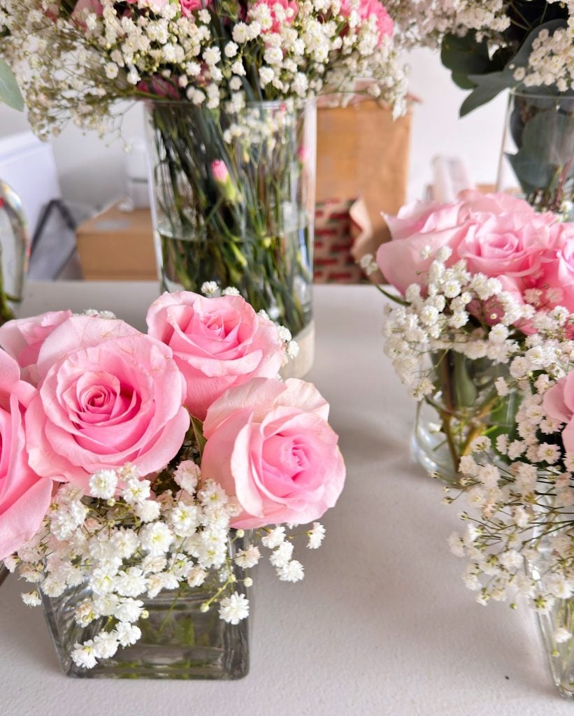 Pink roses and white baby's breath flowers arranged in clear glass vases on a white surface, with more floral arrangements in the background.