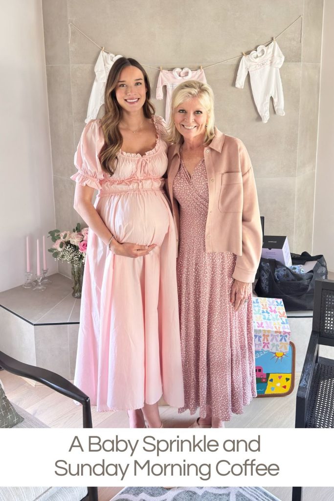 Two women stand side by side indoors, both wearing pink dresses. One is pregnant and holding her belly. Behind them are baby clothes on a line and wrapped gifts on the floor.