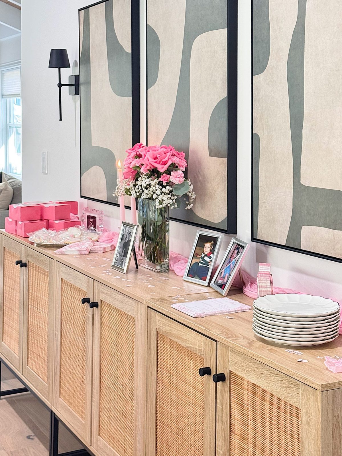 A wooden sideboard displays pink gift boxes, plates, framed photos, flowers in vases, and a lit candle, set against abstract art on the wall.