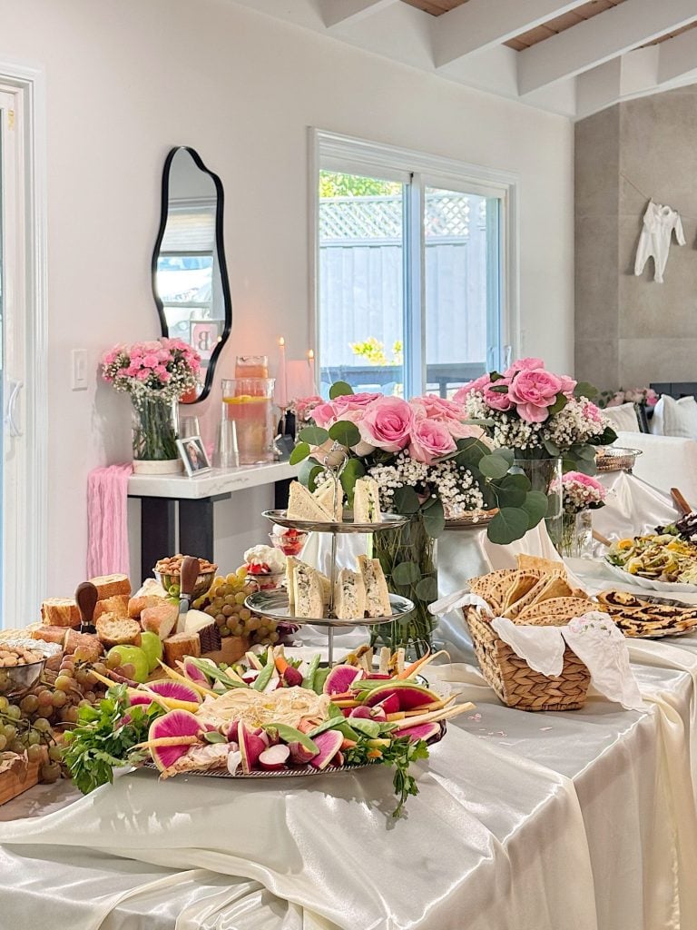 A table set with assorted cheeses, fruits, breads, and vegetables on white satin, with pink flowers and candles in the background near a window.