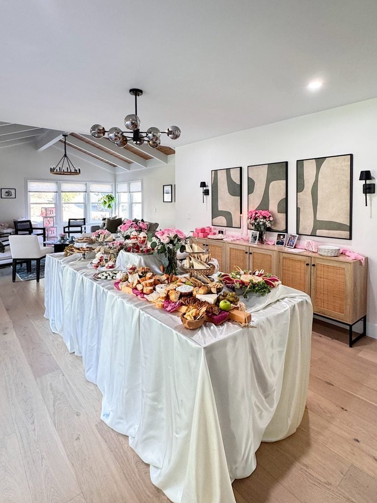 A long table covered with a white tablecloth is set with assorted pastries, flowers, and decorations in a modern, well-lit living room.