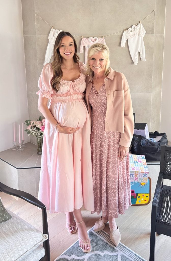 Two women stand indoors, both wearing pink dresses. One is visibly pregnant. Baby clothes hang on a line behind them and there are gifts and flowers on a table nearby.
