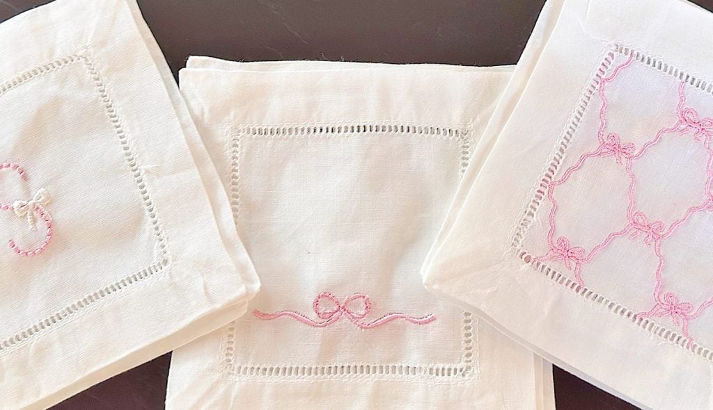 Three white linen napkins with pink embroidered designs and decorative borders are arranged on a dark surface.