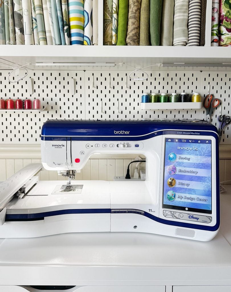 A Brother Innov-is sewing and embroidery machine is on a white table, with fabric, thread spools, and sewing supplies organized on shelves behind it.