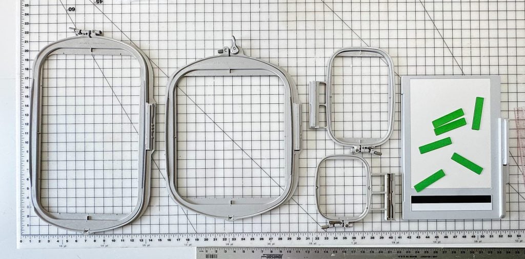 Five metal embroidery hoops of various shapes and sizes are arranged on a gridded cutting mat next to a white rectangle with green tape pieces and a black stripe.