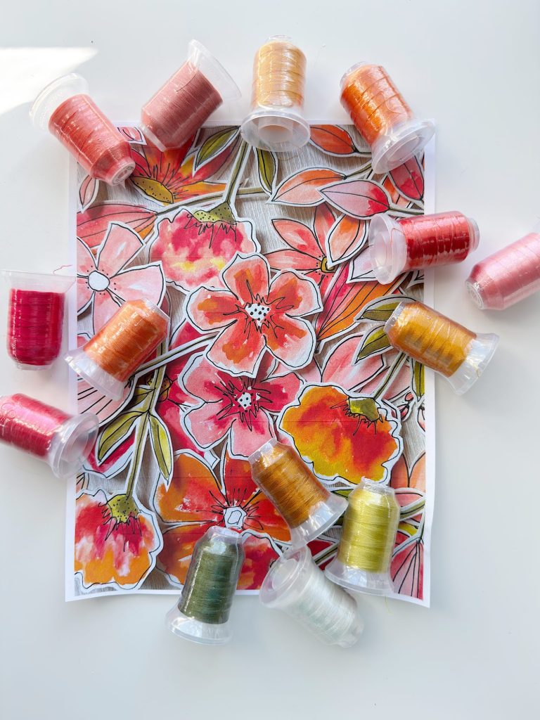 A sheet with a colorful floral drawing is surrounded by spools of thread in various shades of red, orange, yellow, pink, and green on a white surface.