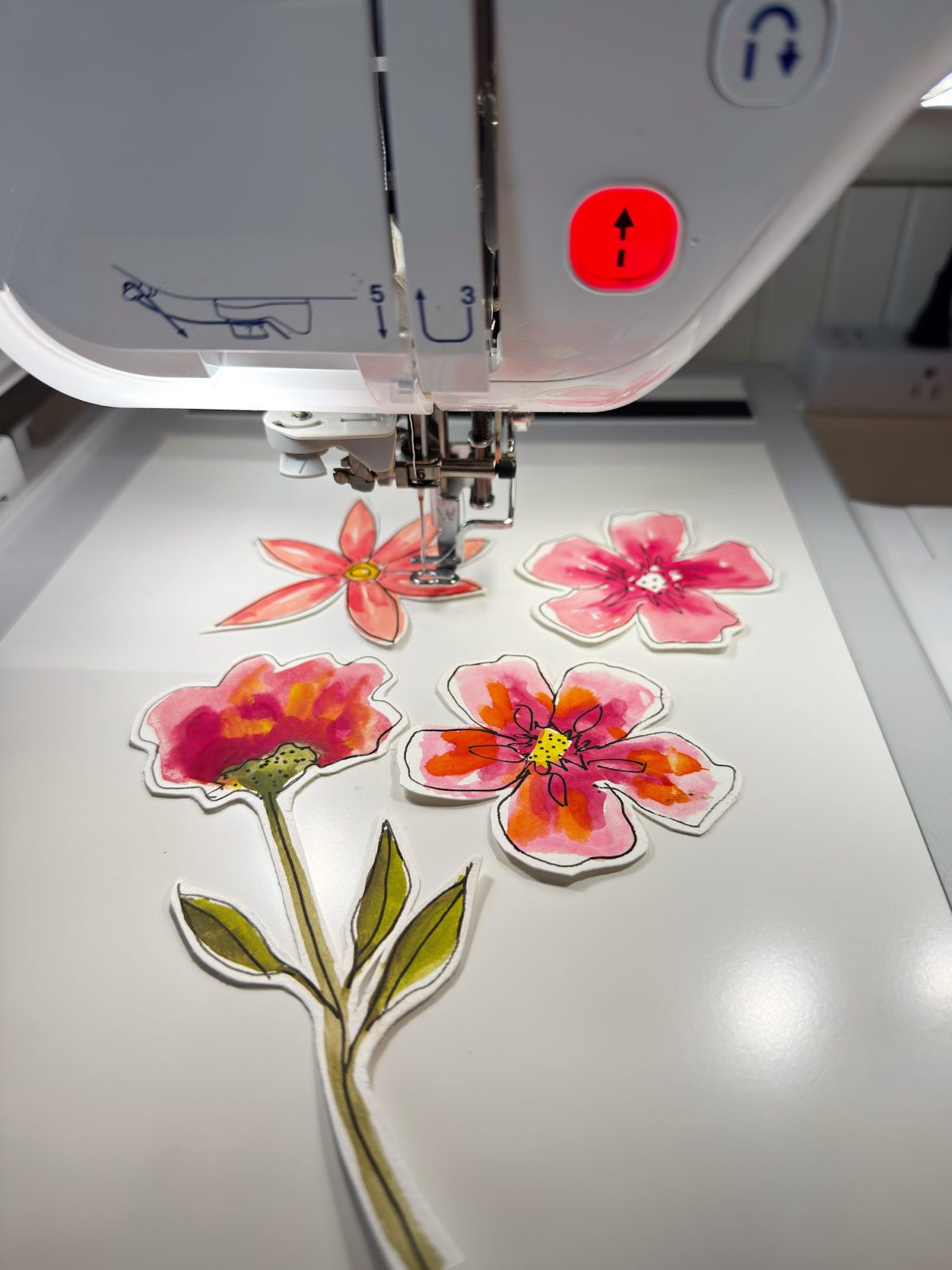 A sewing machine is stitching colorful fabric flower appliqués with pink and red petals onto a white surface.