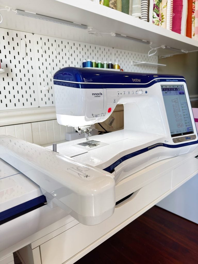 A computerized Brother Innov-is sewing and embroidery machine sits on a white table in a well-organized sewing room with thread spools and fabric on shelves.
