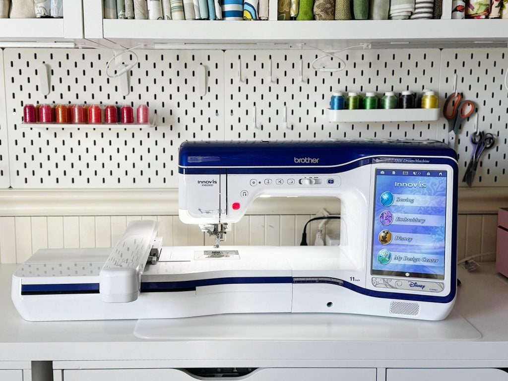 A Brother embroidery and sewing machine with a digital touch screen sits on a white desk, surrounded by shelves holding colorful spools of thread and sewing supplies.
