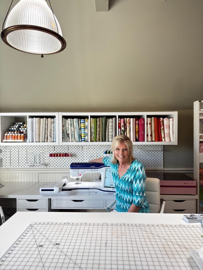 A woman sits at a sewing machine in a well-organized craft room with shelves of fabric, thread, and supplies in the background.