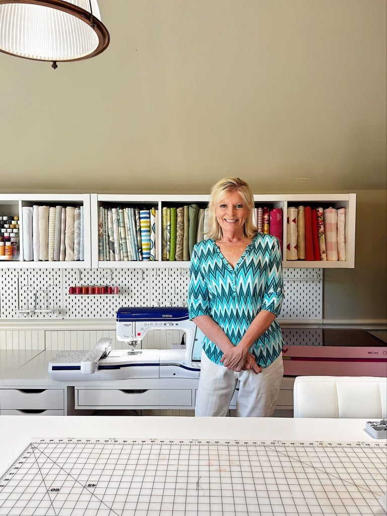 A woman stands and smiles in a craft room, with shelves of fabric, a sewing machine, and a large cutting mat visible in the background.