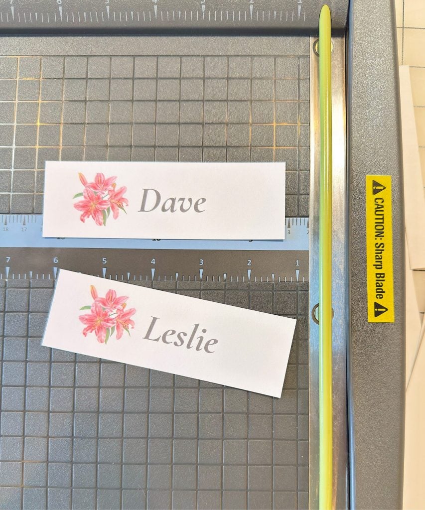 Two name cards labeled "Dave" and "Leslie" with pink flowers are placed on a paper cutter with a "Caution: Sharp Blade" warning label.