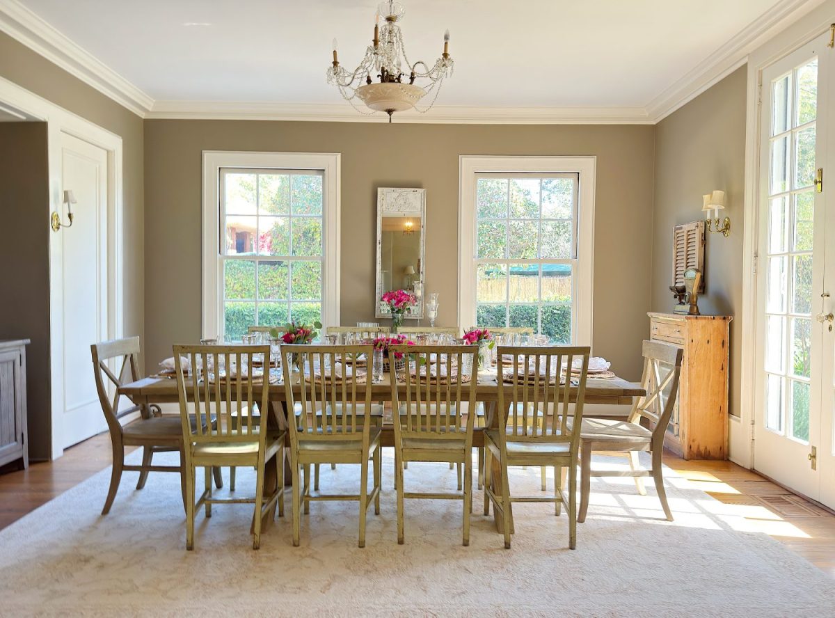 A bright dining room with a large wooden table, ten chairs, chandelier, and flowers on the table, featuring three tall windows and a sideboard.
