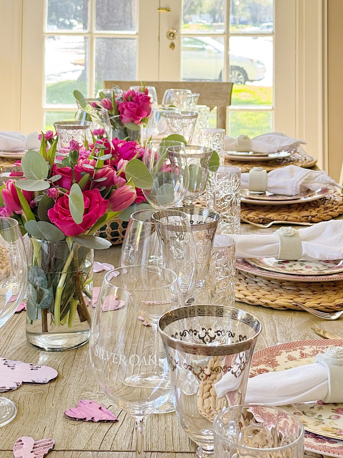 A dining table set with floral arrangements, glassware, plates, and napkins, with sunlight streaming through large windows in the background.