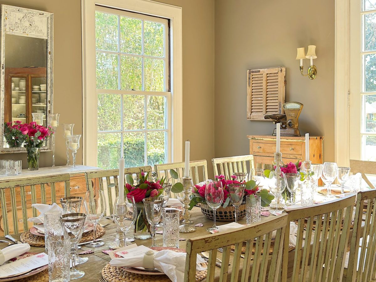A dining room with a long wooden table set for a meal, featuring pink floral centerpieces, glassware, and natural light from large windows.