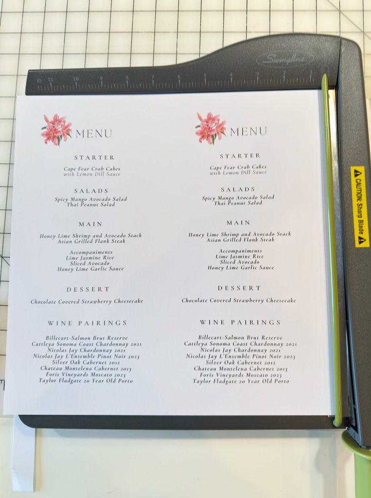 Two printed menus with floral accents list options for starter, salads, main, dessert, and wine pairings on a cutting board with a paper trimmer beside them.