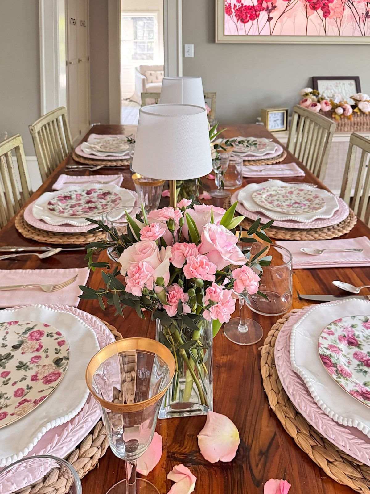 A wooden dining table set with floral plates, glassware, pink napkins, a lamp, and a vase of pink flowers, with rose petals scattered around.