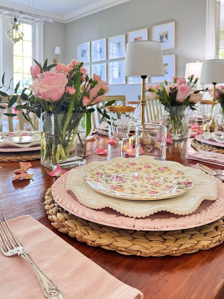 A dining table set with floral plates, pink napkins, woven placemats, glassware, and vases of pink flowers in a bright, elegant room.