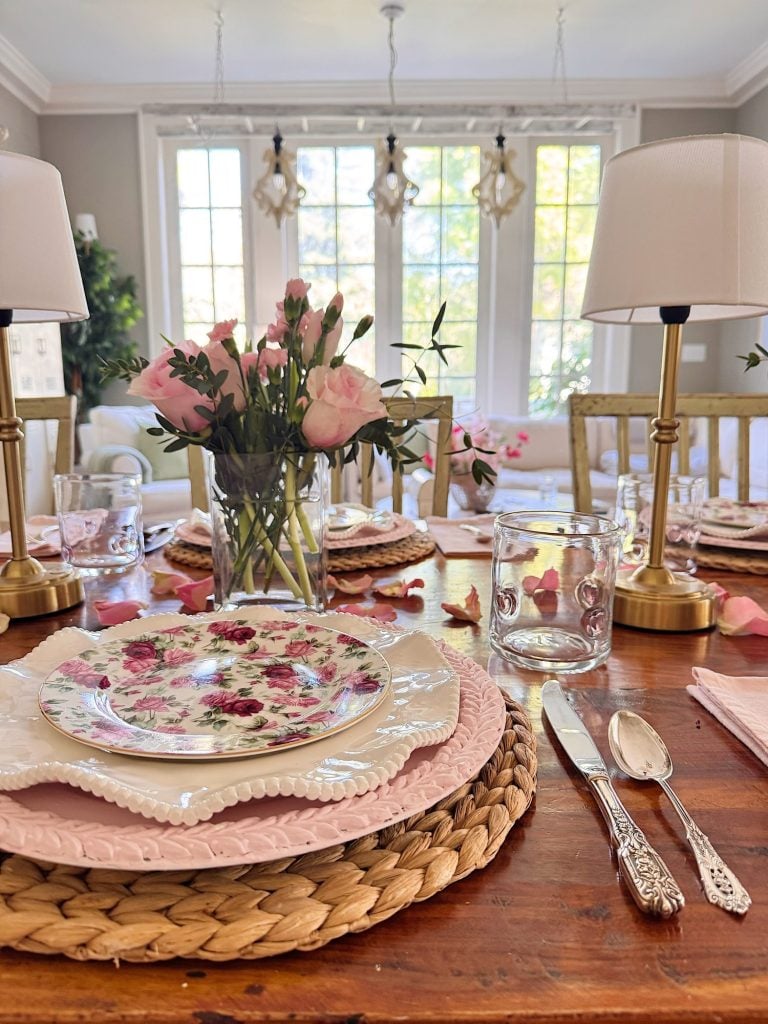 A dining table set with floral plates, pink napkins, silverware, a vase of pink flowers, two lamps, and rose petals, in a bright room with large windows.