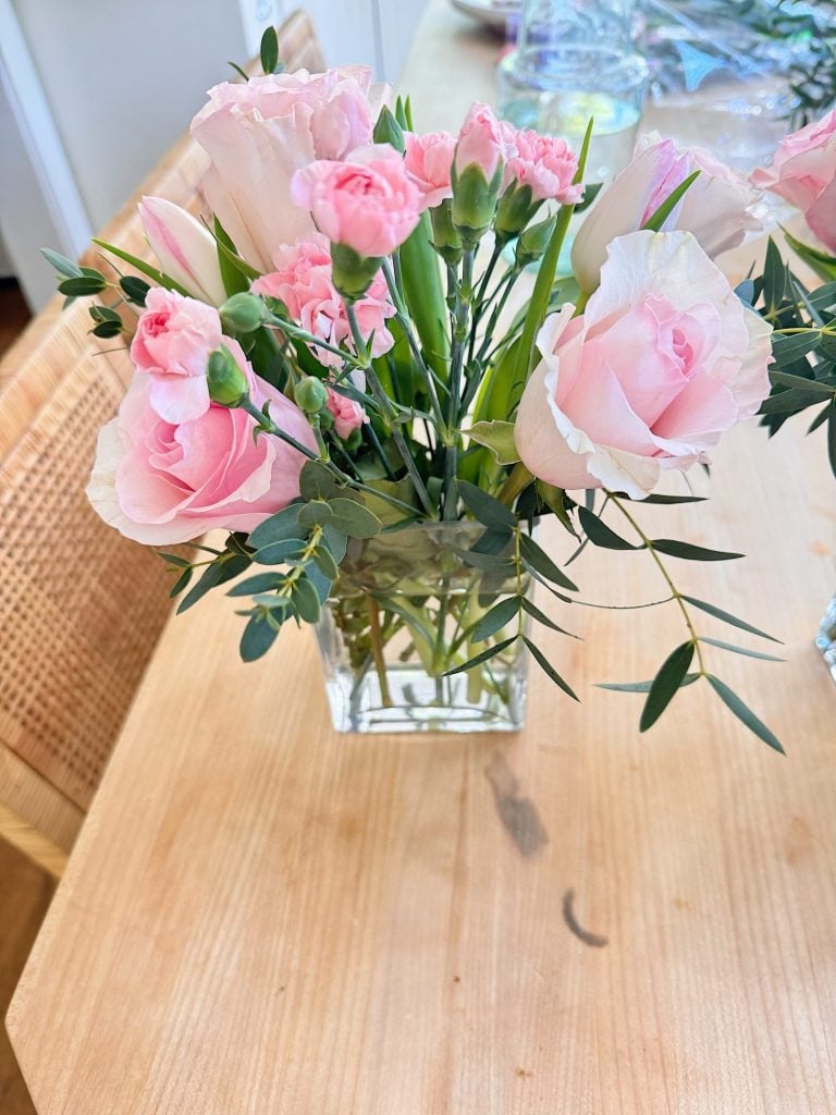 A small glass vase with pale pink roses, pink carnations, and green foliage sits on a light wooden table.