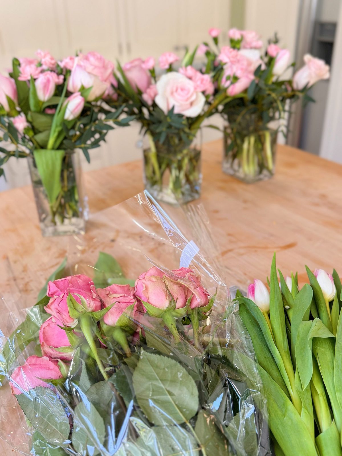 Four glass vases filled with pink roses and tulips are on a wooden table. In the foreground, pink roses in plastic wrap and green tulip stems are visible.