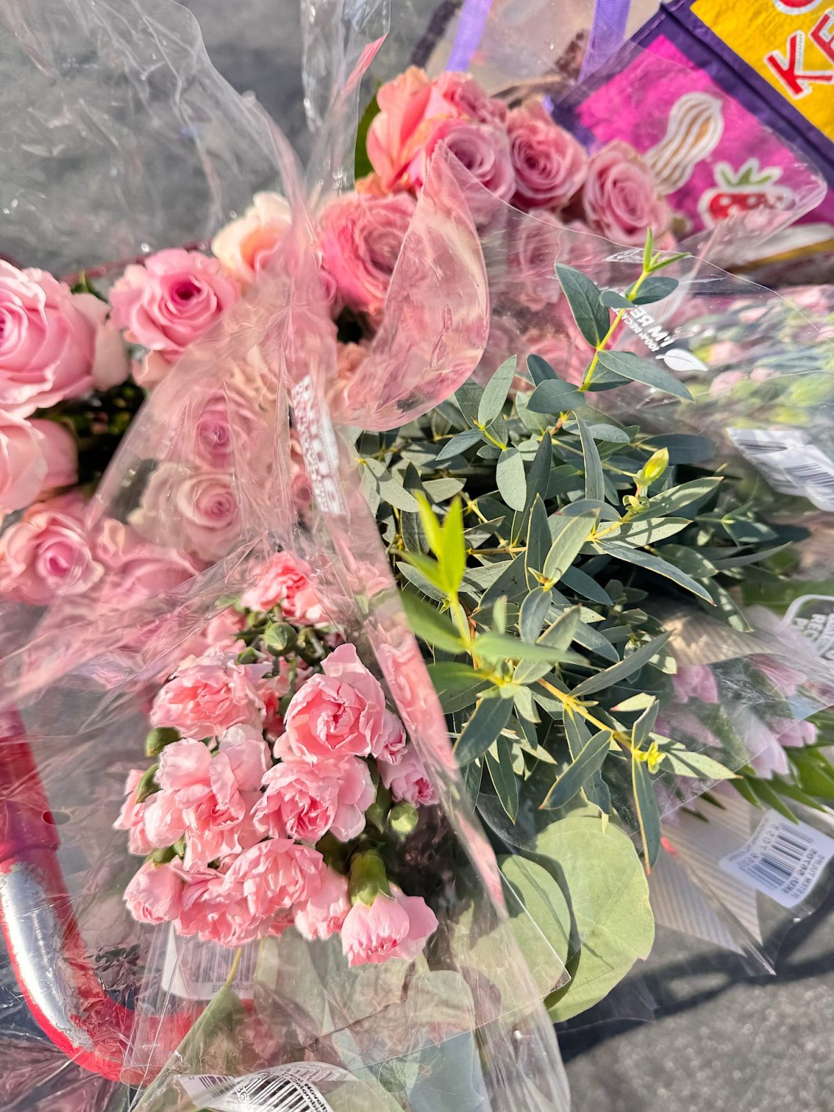 Bouquets of pink flowers and green foliage are wrapped in plastic and placed in a shopping cart, partially visible with a package of Kellogg’s Frosted Strawberry Pop-Tarts in the background.
