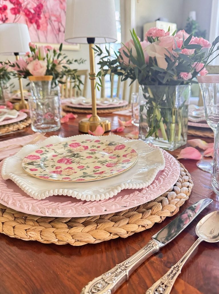 A close-up of a table set with floral plates, woven placemats, silverware, pink flowers in vases, and glassware, suggesting a decorative dining setting.