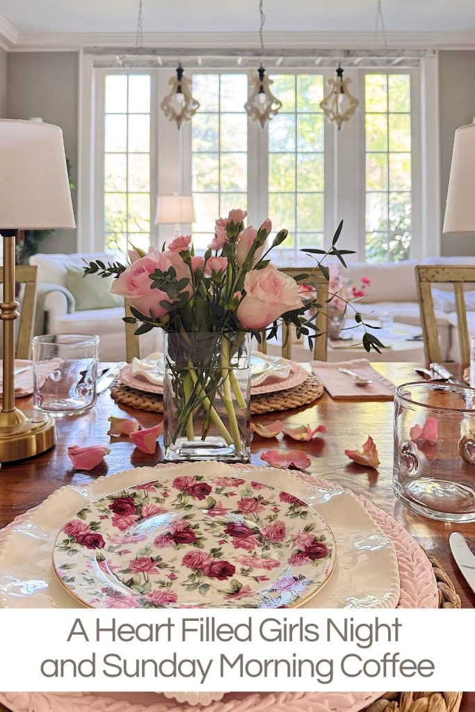 A dining table set with floral plates, pink flowers in a vase, glasses, and scattered rose petals; a sofa and large windows in the background.