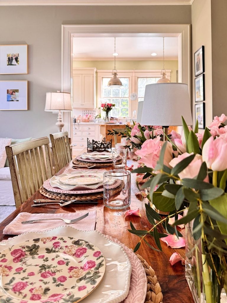 A wooden dining table set for a meal with floral plates, glasses, cutlery, and pink flowers; a bright kitchen is visible in the background.