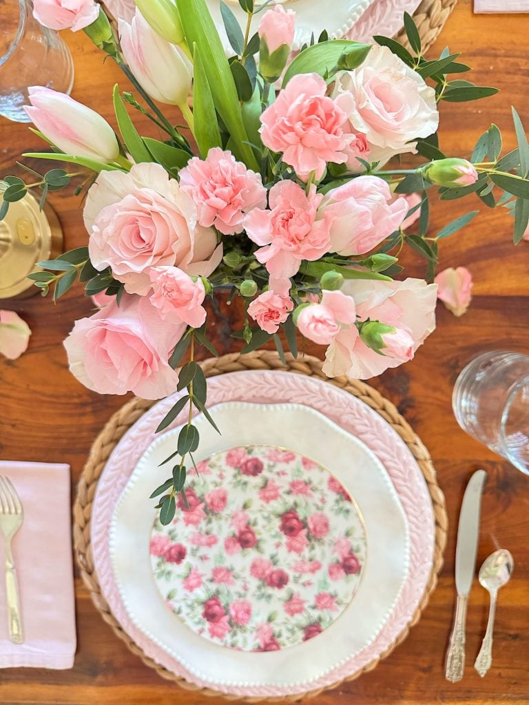 A table setting with a pink floral plate, white dishes, silver cutlery, pink napkin, and a centerpiece of pink flowers and greenery on a wooden table.