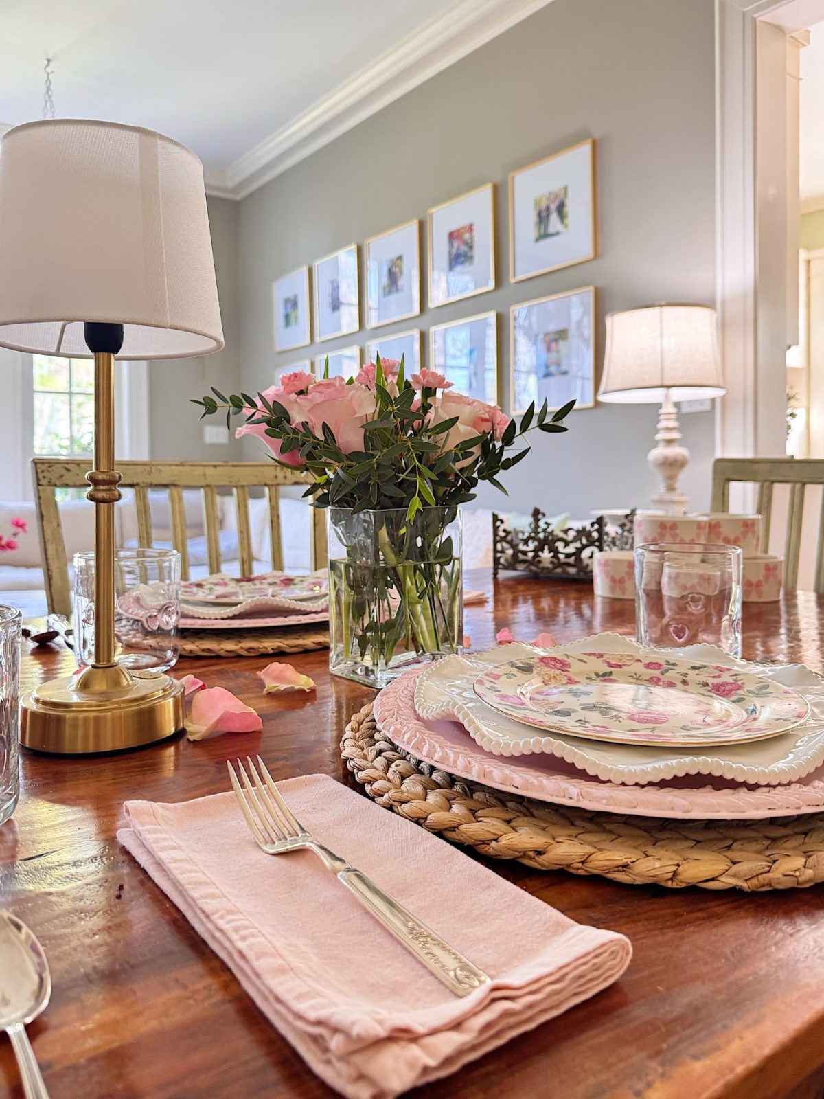 A wooden dining table set with floral plates, pink napkins, a fork, a glass, a vase of pink roses, and decorative lamps, in a well-lit room with framed photos on the wall.
