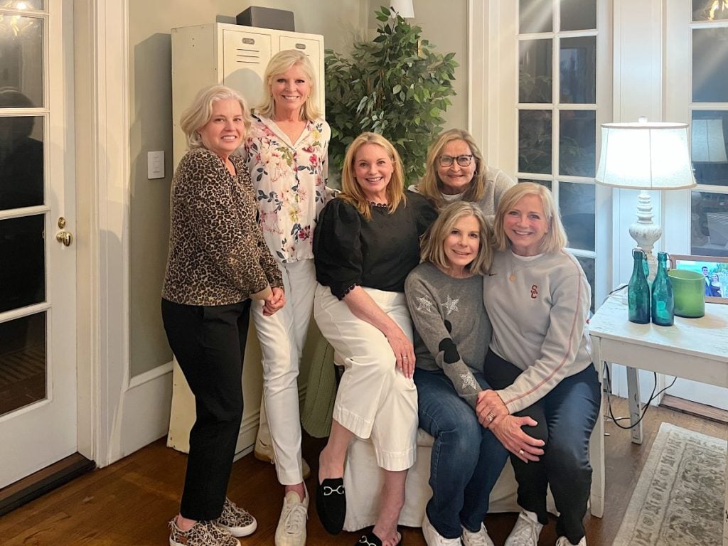 Six women pose together indoors, standing and sitting near a table with bottles, a lamp, and a potted plant, with large windows in the background.
