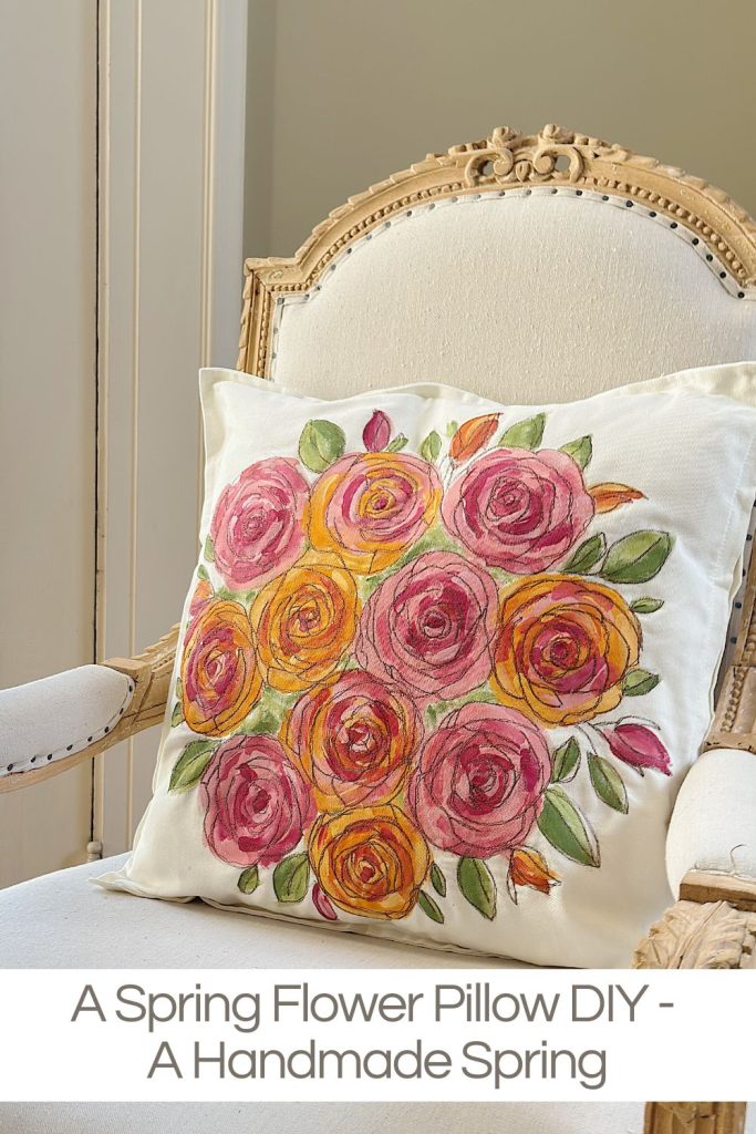 A cream-colored armchair with a white pillow featuring a colorful hand-painted floral design, placed near a window.