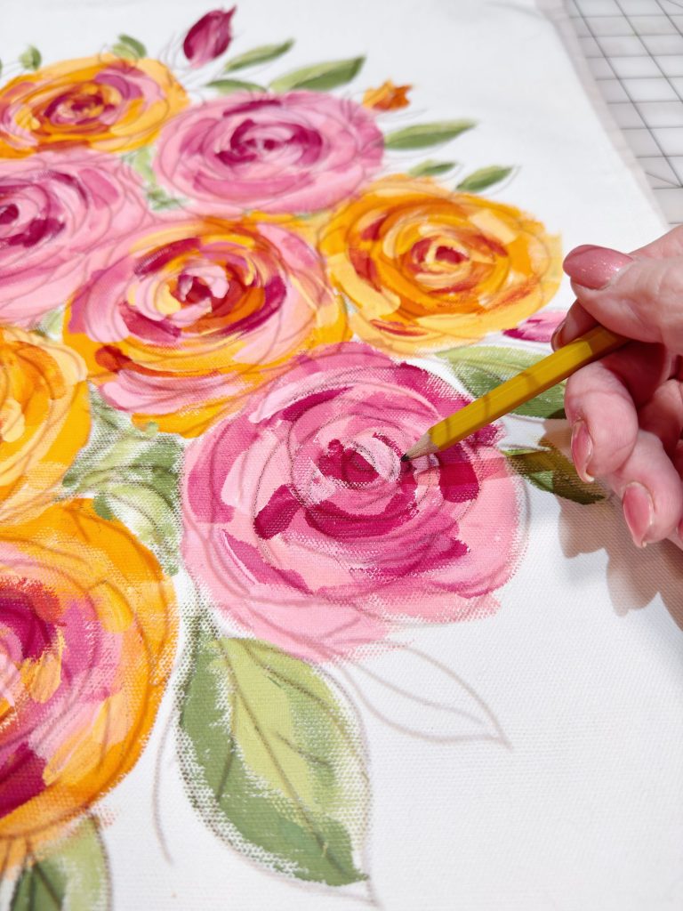 A hand uses a yellow paintbrush to add details to pink and yellow roses painted on white canvas with green leaves.