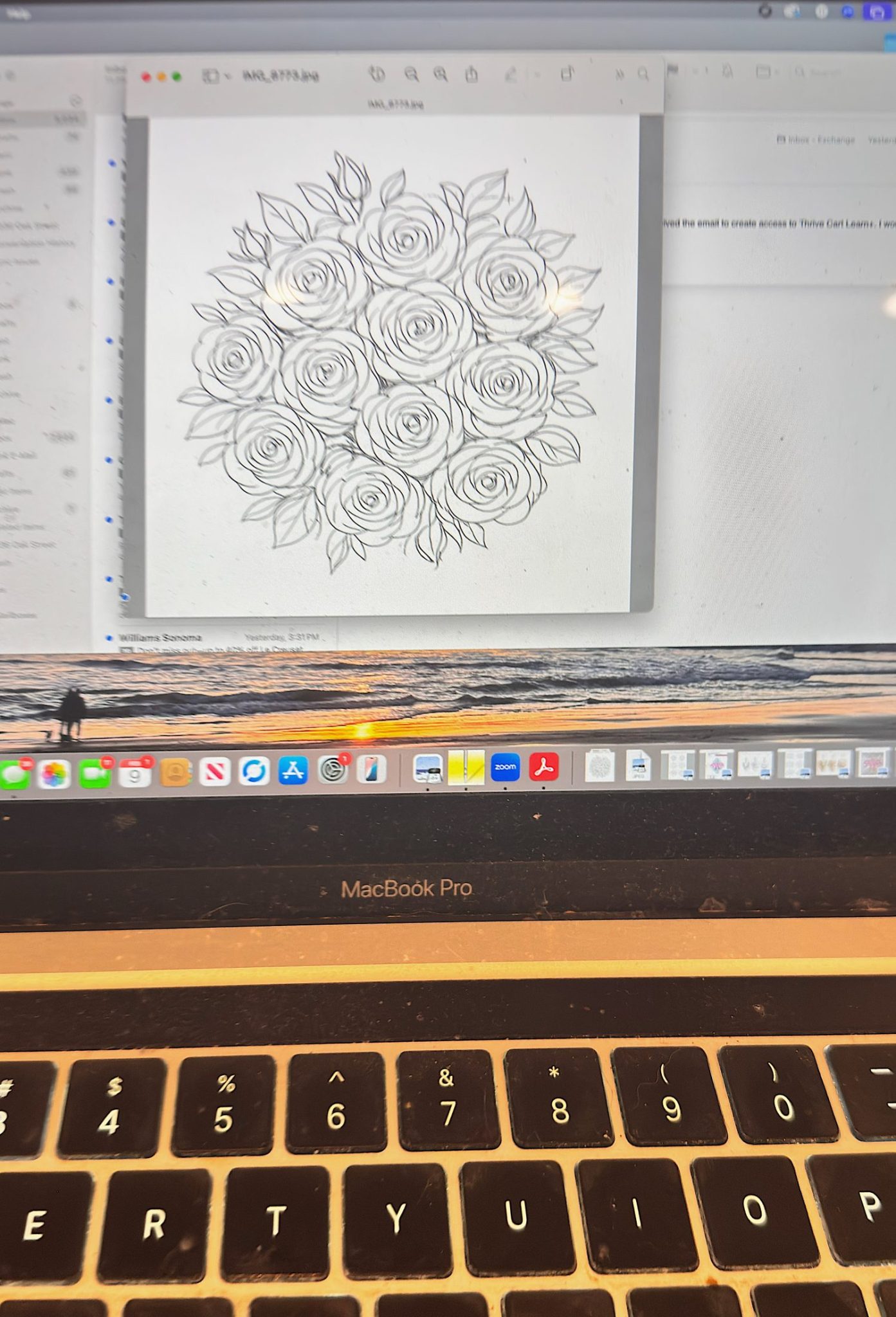 A MacBook Pro displaying a black-and-white floral drawing on the screen, with a beach-themed desktop wallpaper partially visible in the background.