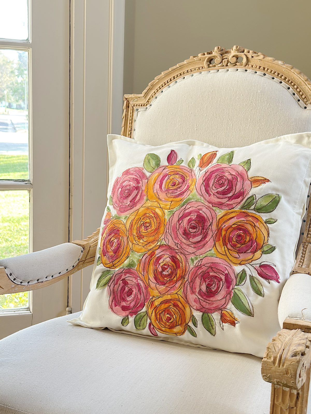 A white upholstered armchair with a pillow featuring a colorful painted floral design in pink, orange, and green, set near a window with sunlight streaming in.
