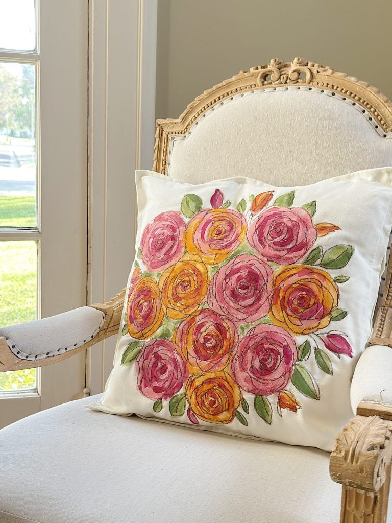 An ornate cream armchair with a white cushion featuring a colorful floral design of pink and yellow roses with green leaves, placed near a window.