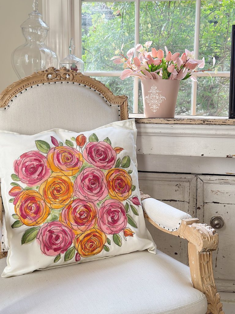 An upholstered chair with a floral pillow sits next to a white cabinet and a window; a pink flower arrangement is on the cabinet.