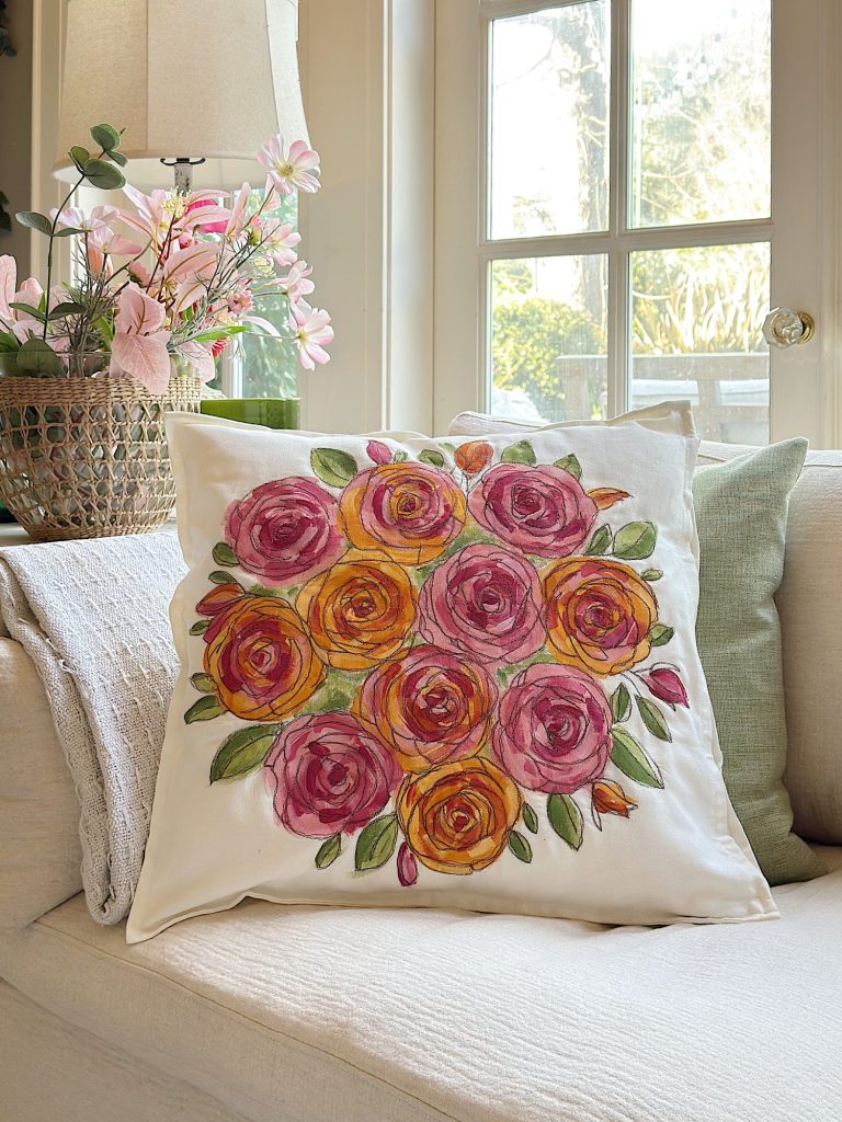 A white throw pillow with a colorful rose bouquet design sits on a cream sofa, next to green pillows, with a vase of pink flowers and a window in the background.