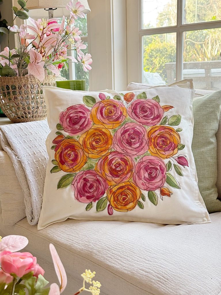 A white throw pillow with a painted floral design of pink and yellow roses sits on a cream-colored sofa in a bright living room.