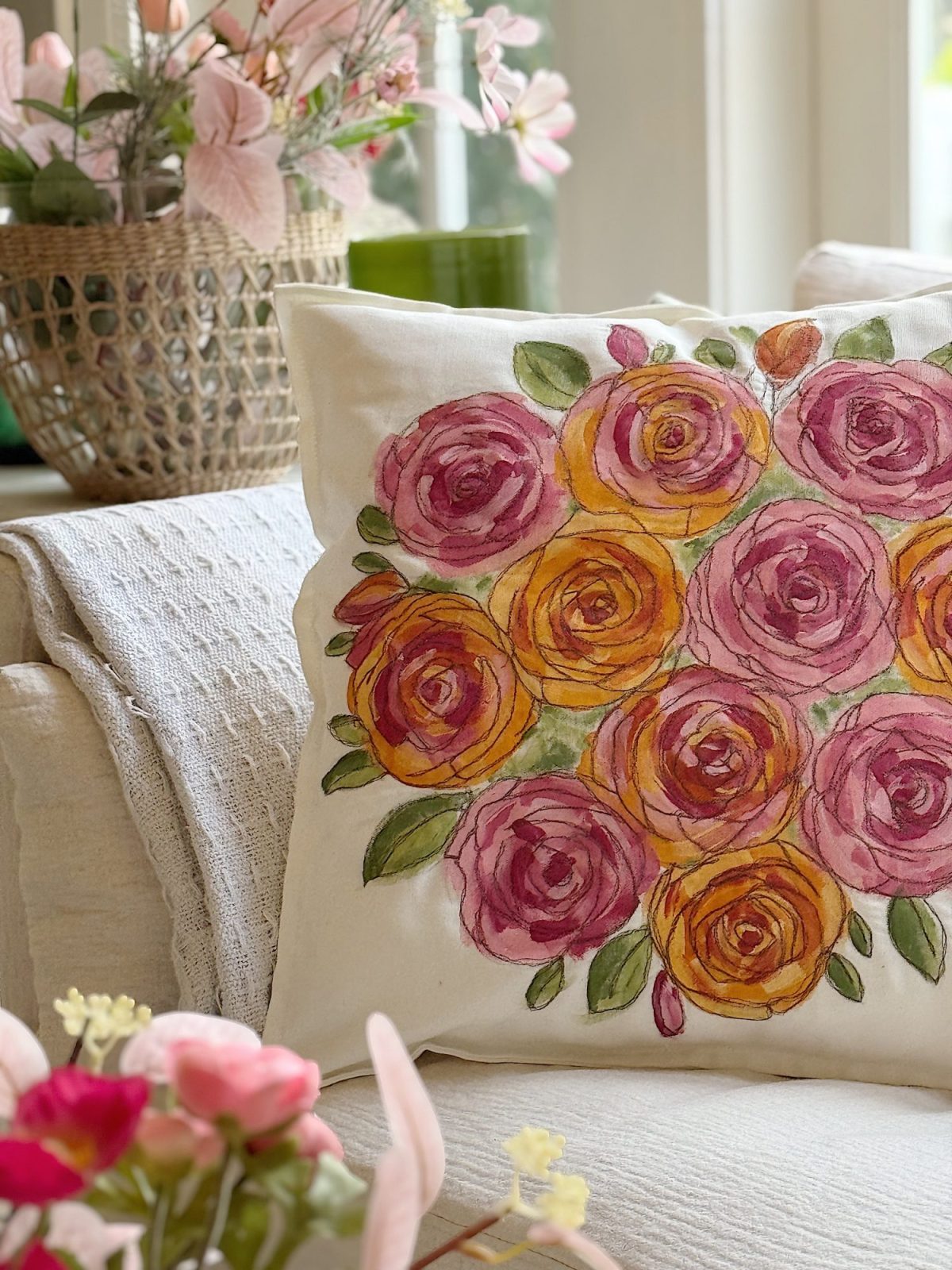 A decorative pillow with a watercolor-style rose pattern in pink and orange rests on a cream-colored couch, surrounded by flower arrangements.