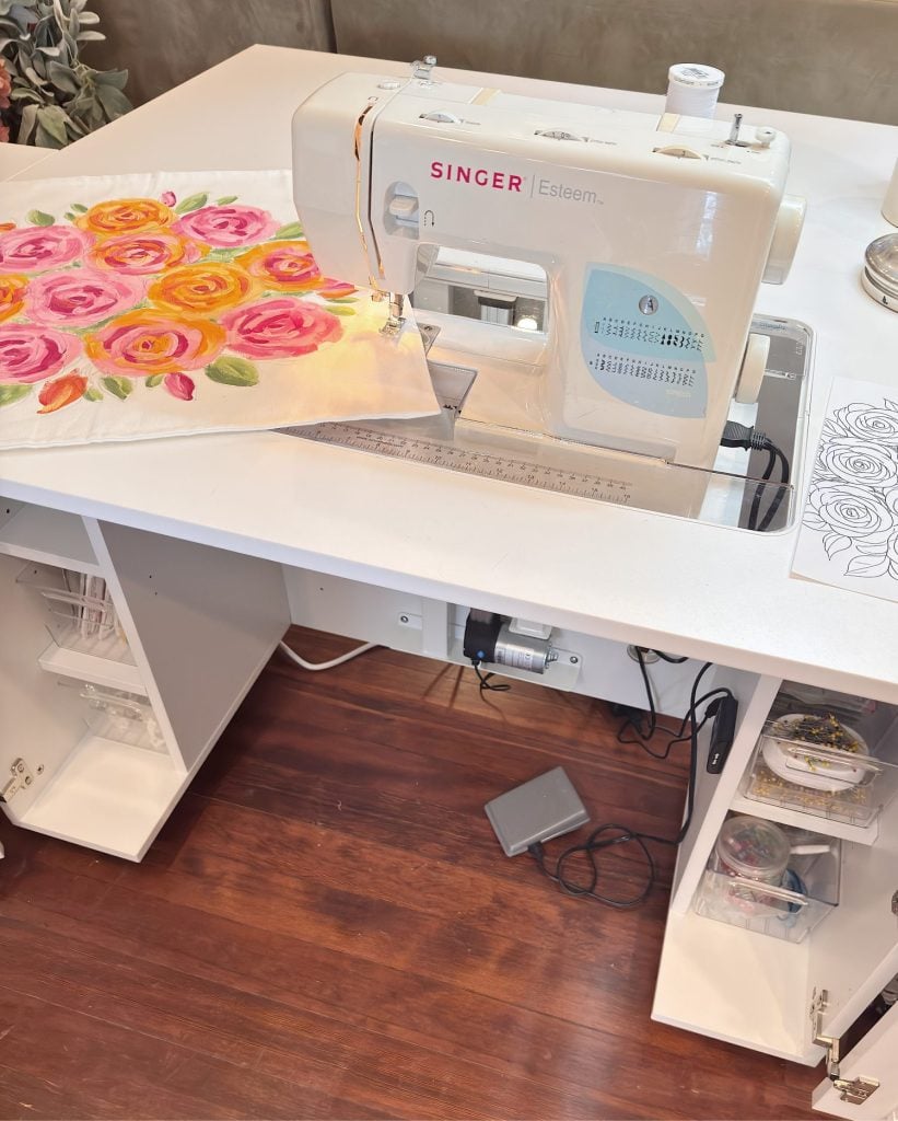 A Singer Esteem sewing machine on a white desk with a floral fabric piece being stitched; sewing supplies and tools are stored on shelves below the desk.