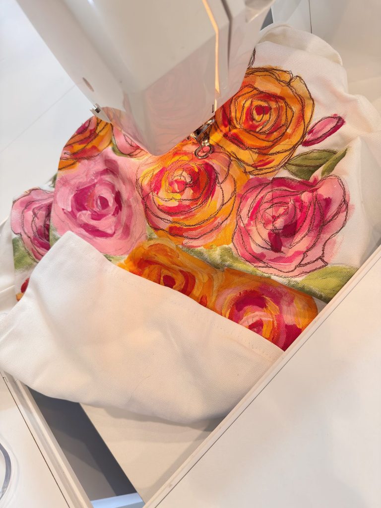 A sewing machine stitches a piece of white fabric with colorful, hand-painted rose designs in pink and orange hues.